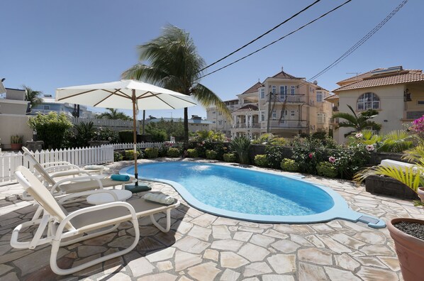 Une piscine entourée d’une terrasse en pierre et d’un aménagement paysager luxuriant, avec des chaises longues et un parasol à proximité.