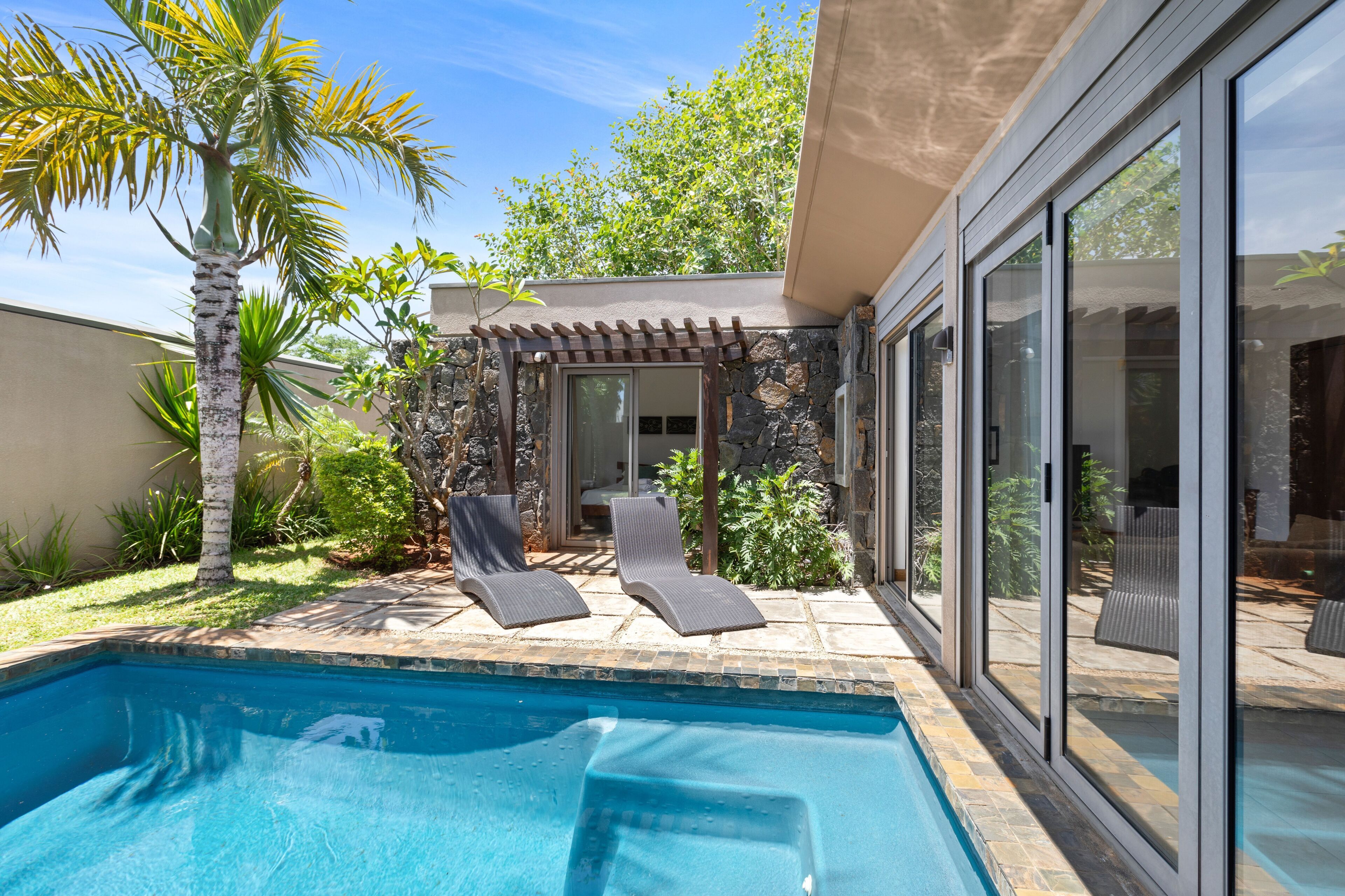 A swimming pool with lounge chairs, a pergola, and a stone wall.