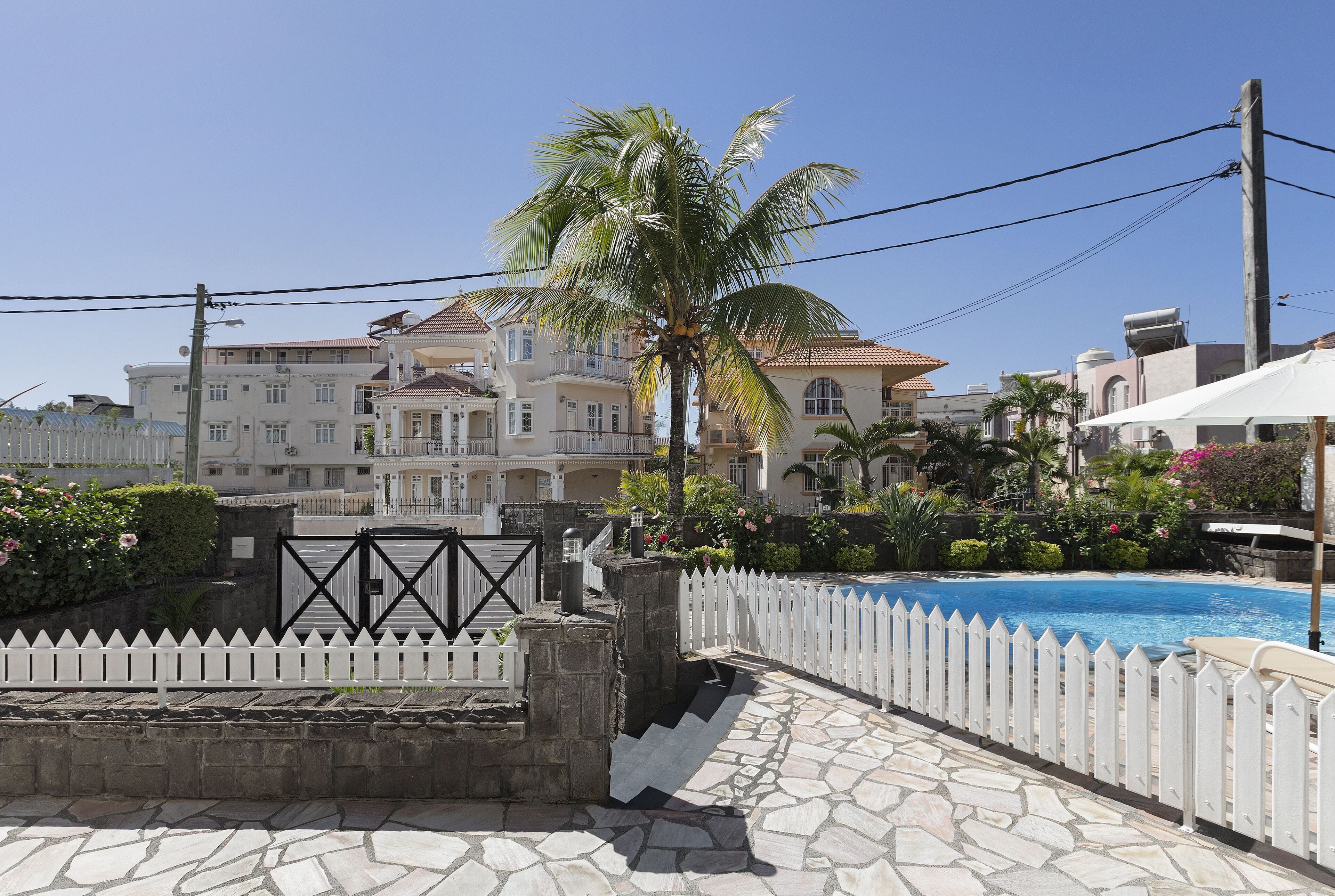 A pool area with a white picket fence, a palm tree, and a stone pathway leading to a gated entrance.