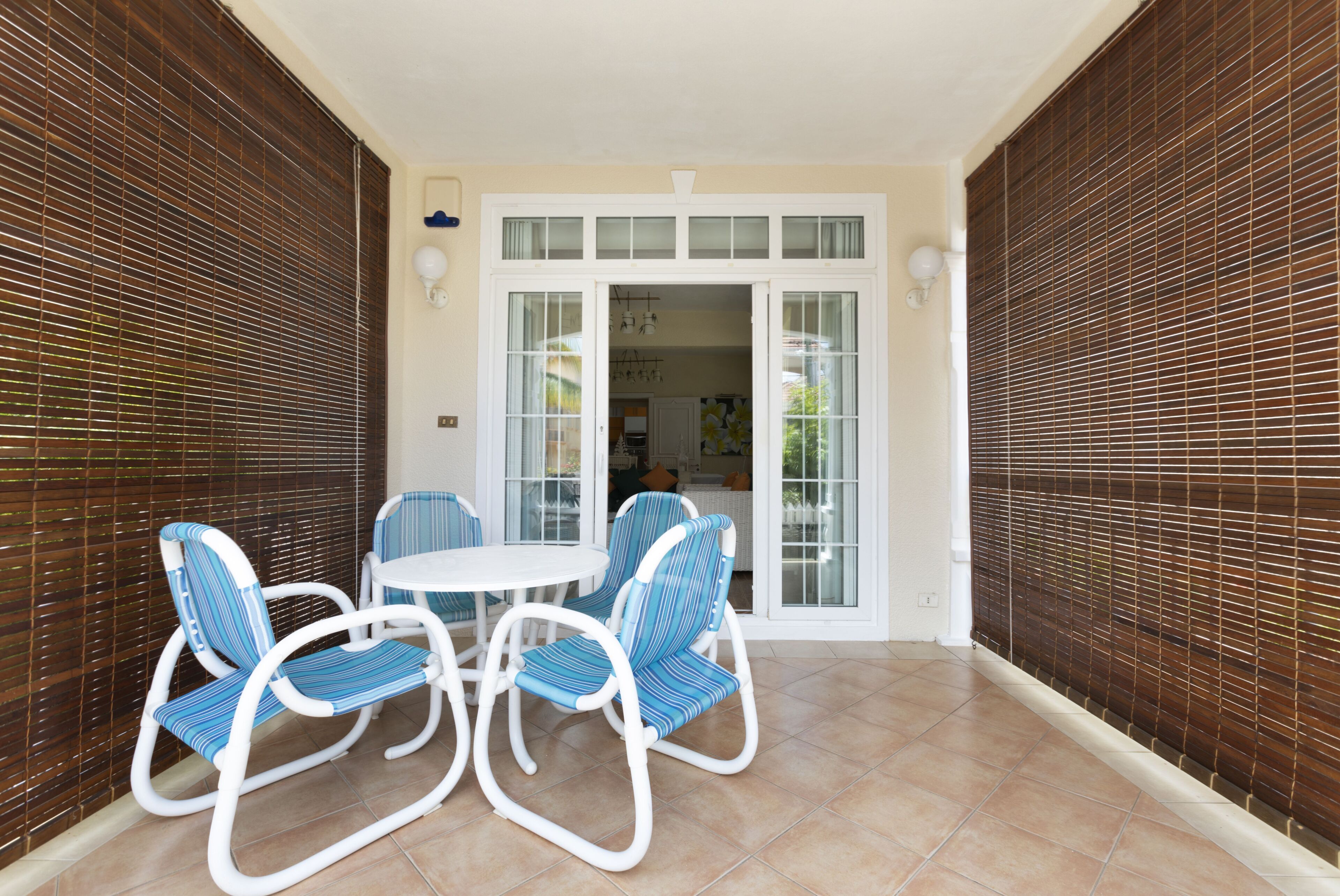 Ein überdachter Patio-Bereich mit einem runden Tisch und vier gestreiften Stühlen, einer weißen Decke und Blick in die Küche durch eine Glastür.