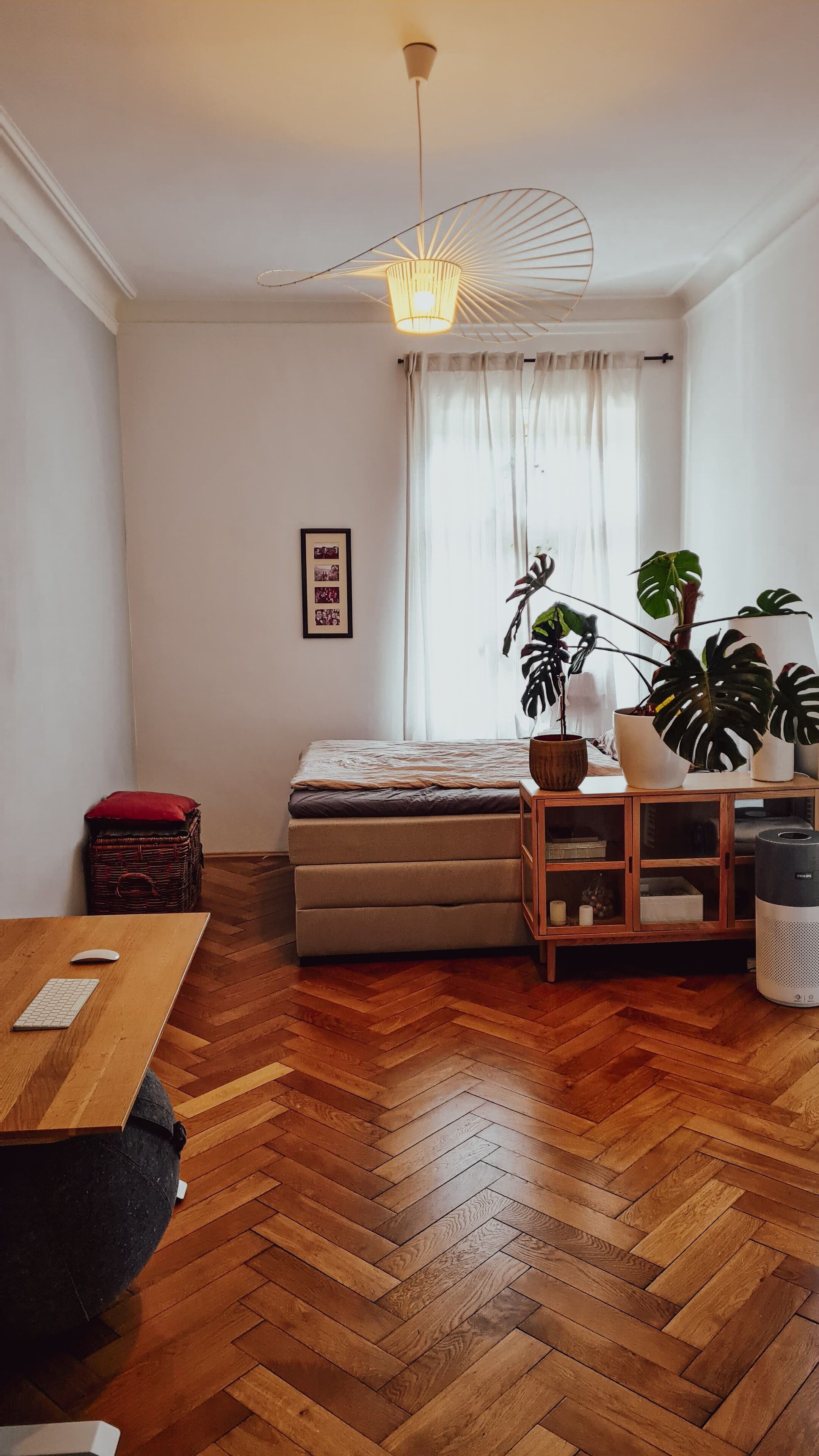 A modern living room with a wooden herringbone floor, a sofa, a wooden coffee table, a hanging light fixture, and a potted plant.