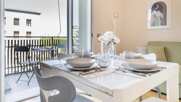 A dining area with a table set for two, a balcony with a view of buildings, and a framed picture on the wall. - Easylife - View and confort in Porta Vittoria (Milano)