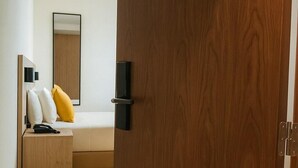 Een moderne slaapkamer met een houten deur, een bed met witte en gele kussens, een nachtkastje met een telefoon en een spiegel aan de muur.