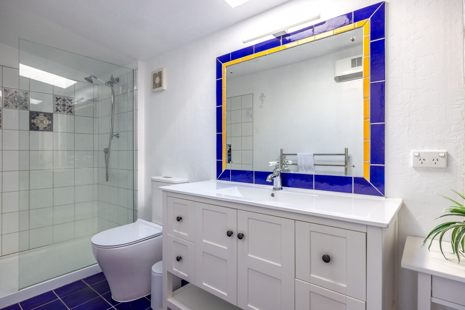 Une salle de bain dotée d’une douche à parois de verre, d’un meuble-lavabo blanc et d’un miroir orné d’une bordure de carreaux bleus et jaunes.