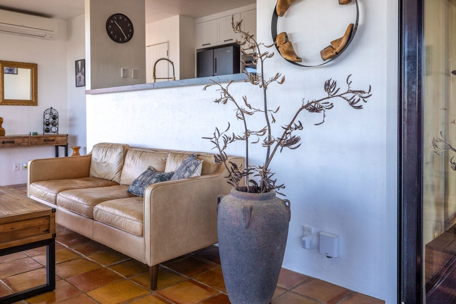 A living room with a beige sofa, a large vase with branches, a clock, and a kitchen area in the background.