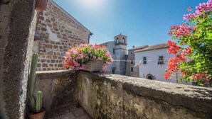 Un muro de piedra con un cactus y flores rosadas, un edificio con una torre de reloj, y un cielo azul despejado.