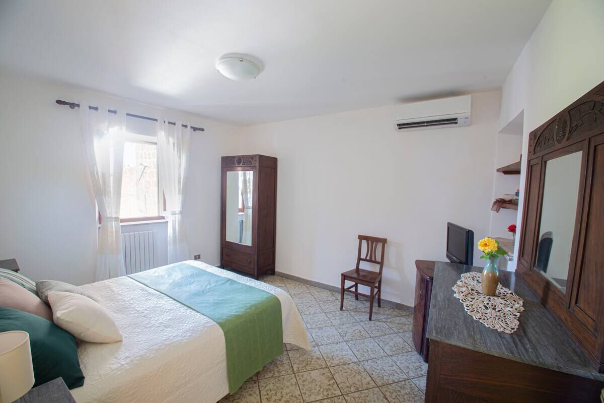 Une chambre à coucher avec un lit, une chaise, une commode, un miroir et une fenêtre avec des rideaux.