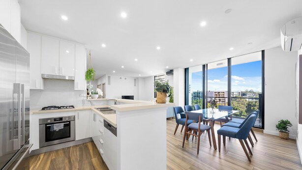 A modern kitchen with white cabinetry, a central island, and a dining area with blue chairs. - Modern 3BR Apt w Parking near ICC & Darling Harbour (Ultimo)