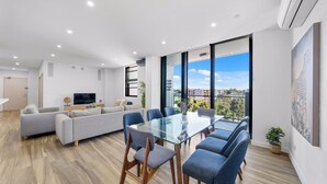 A modern living room with a glass dining table, grey sofas, and a TV. - Modern 3BR Apt w Parking near ICC & Darling Harbour (Ultimo)