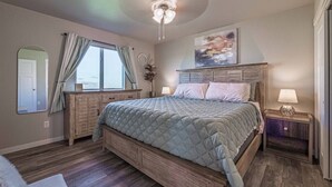 Une chambre à coucher avec un grand lit, une tête de lit en bois, un ventilateur de plafond, une fenêtre avec des rideaux, une table de chevet avec une lampe et un miroir au mur.