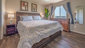 A bedroom with a large bed, wooden floor, a nightstand, a dresser, a ceiling fan, and a window with curtains. - Isaac’s Nook in the Hills (Rapid City)