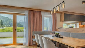 A modern dining area with a wooden table, grey chairs, and a bench. It features large windows with curtains, a view of greenery and mountains. - Newly built chalet in the middle of the Ötztal (Umhausen)