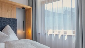 A hotel room with a bed, a wooden headboard, a window with sheer curtains, and a cityscape view. - Newly built chalet in the middle of the Ötztal (Umhausen)