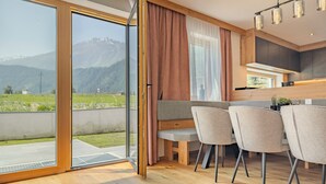A modern kitchen with a wooden countertop, a dining area with chairs, and a sliding glass door leading to a balcony with a mountain view. - Newly built chalet in the middle of the Ötztal (Umhausen)