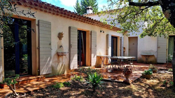 Un bâtiment rustique avec un toit en tuiles, un petit espace extérieur aménagé avec des sièges et un jardin agrémenté de plantes en pot.