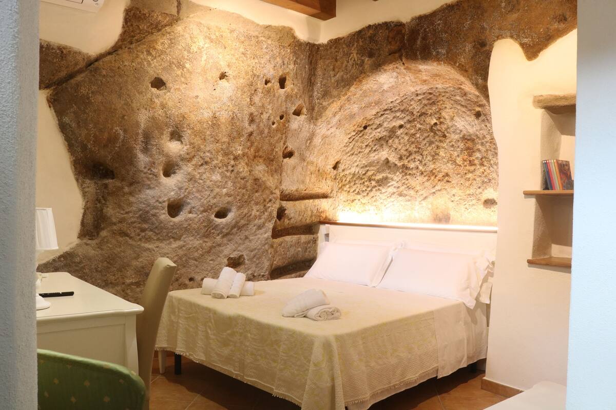 A bedroom with a large rock wall, a bed with white linens, a chair, and a small table with a lamp.