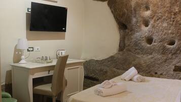 Habitación de hotel con una gran pared de piedra, cama con colcha floral, escritorio con silla, televisor montado en la pared y un pequeño bote de basura verde.
