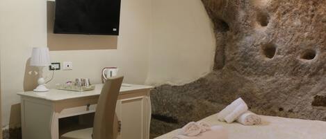 Ein Hotelzimmer mit einer großen Steinwand, einem Bett mit floralem Überwurf, einem Schreibtisch mit Stuhl, einem an der Wand montierten Fernseher und einer kleinen grünen Mülleimer.