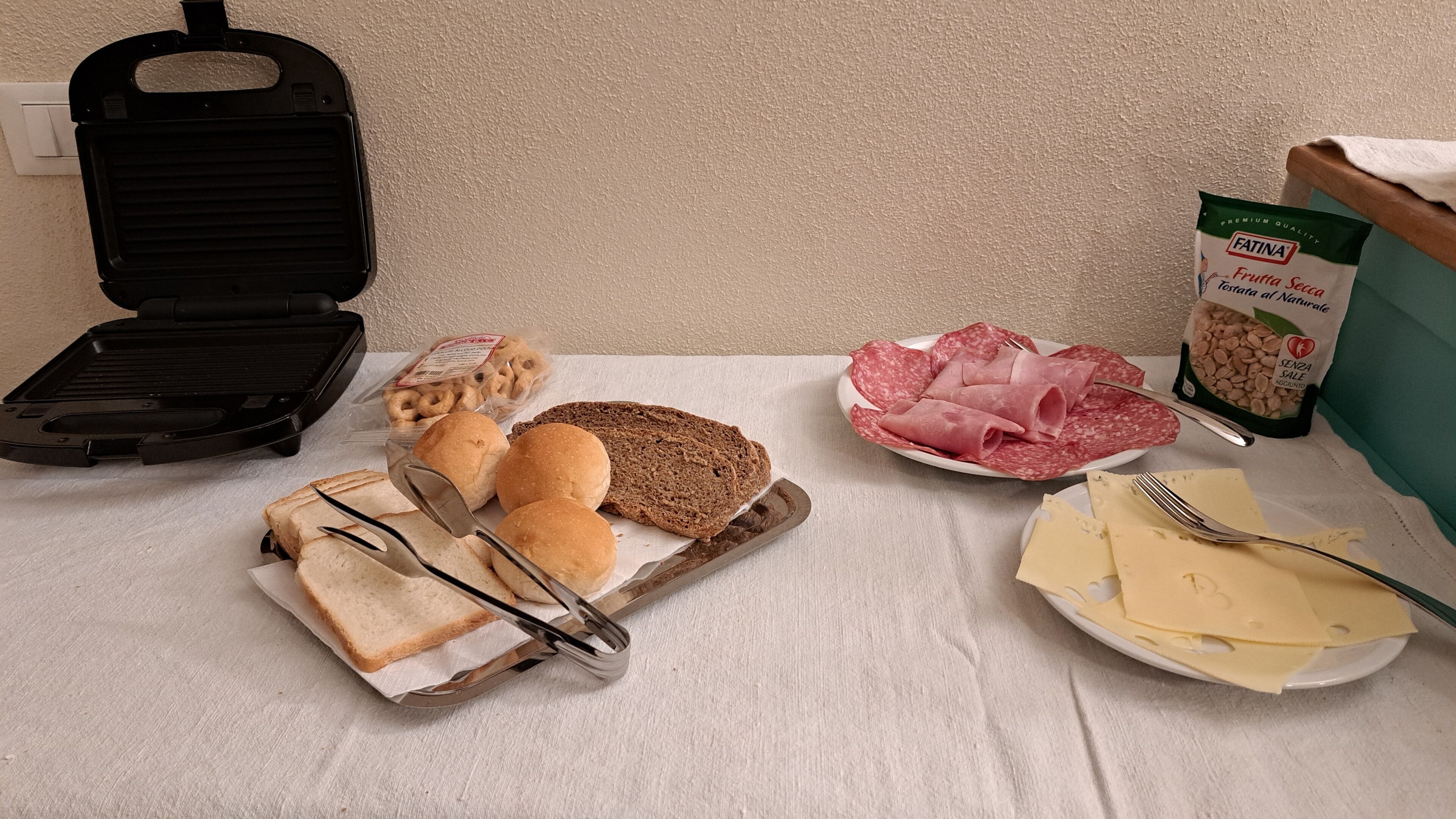 A sandwich board with bread, cheese, and salami, a plate with cheese and a fork, a bag of snacks, and a grill.