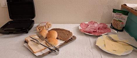 A sandwich board with bread, cheese, and salami, a plate with cheese and a fork, a bag of snacks, and a grill.