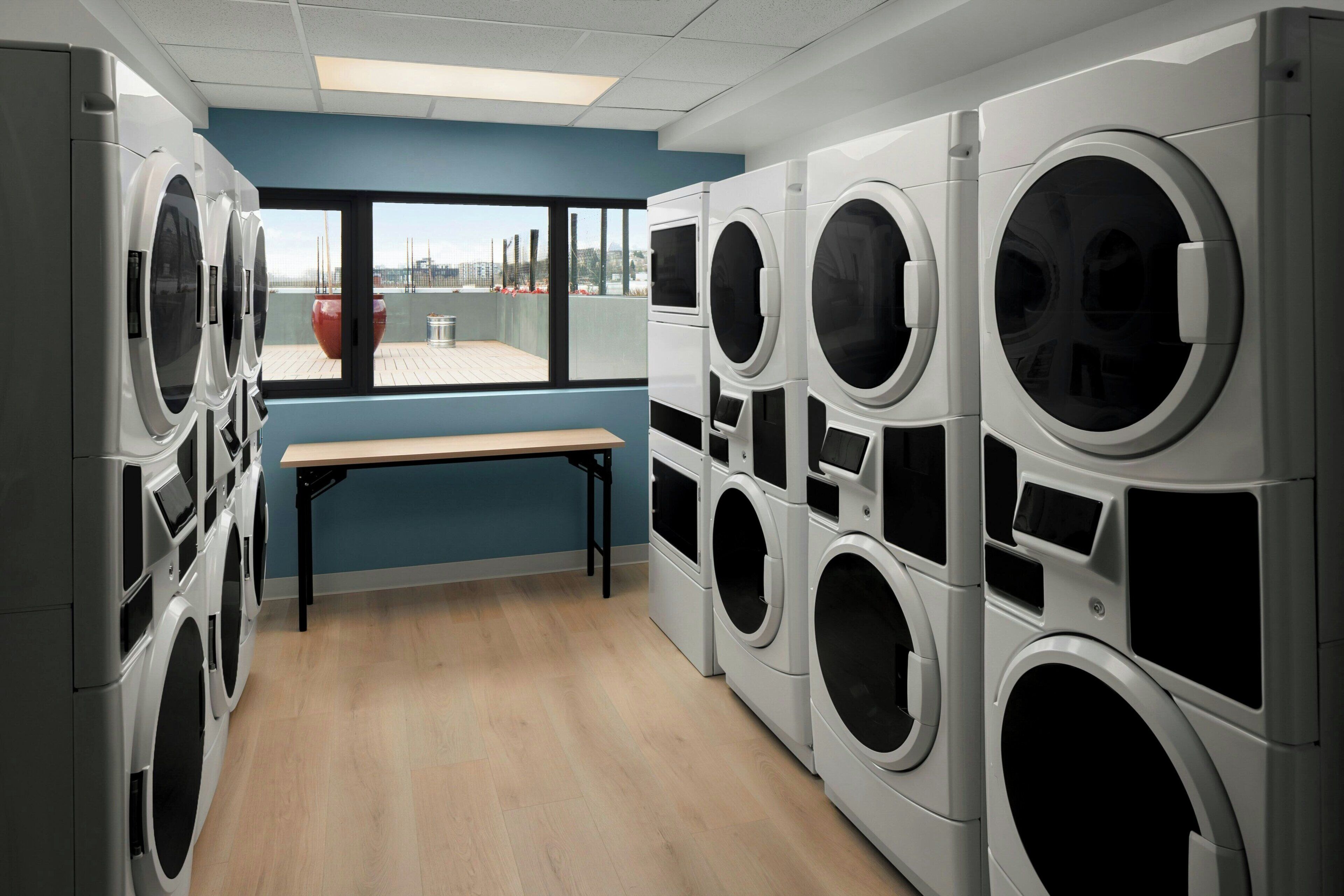 A laundry room with multiple washing machines, a bench, and a large window with a view of buildings.