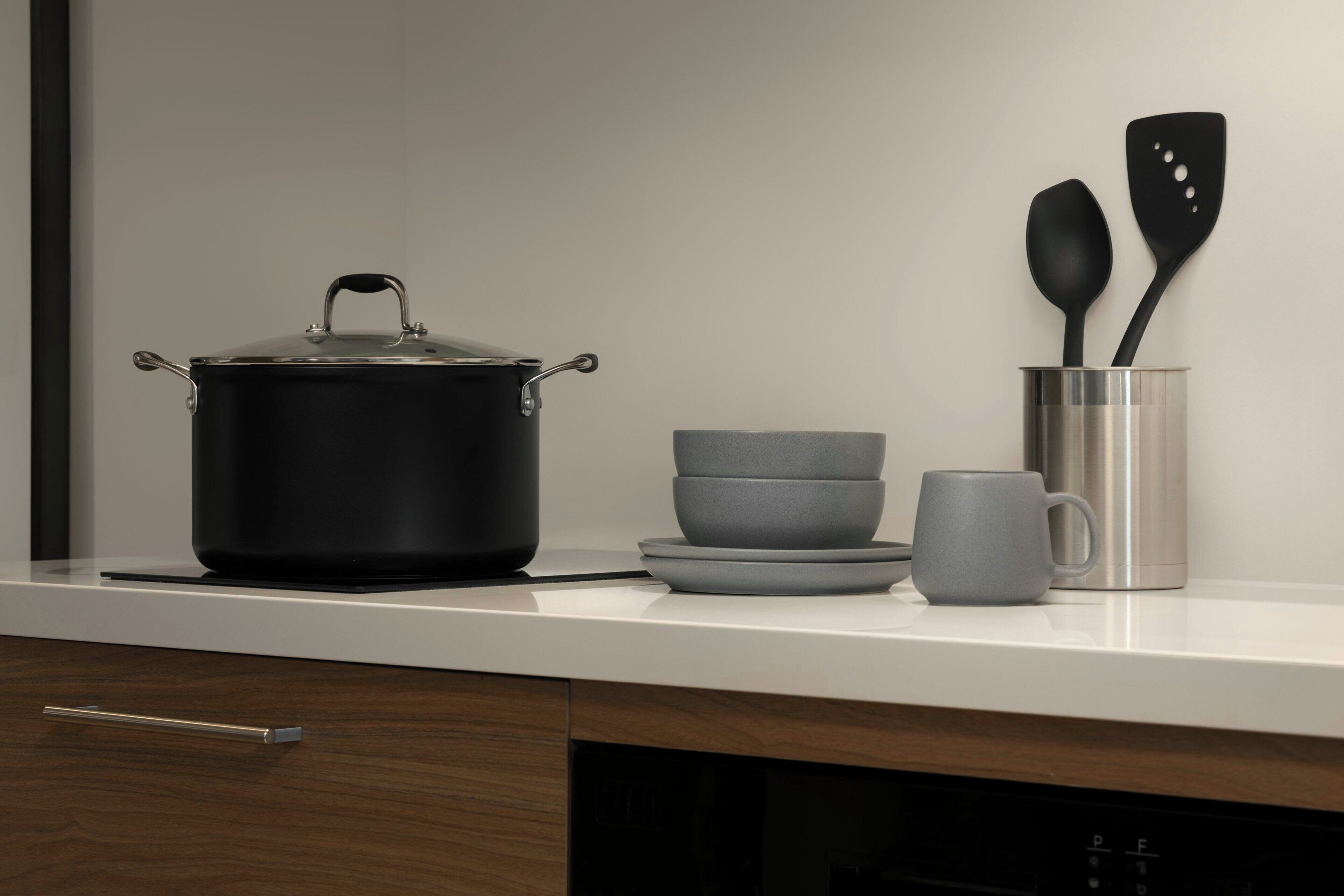 A kitchen counter with a black pot, a stack of gray bowls, a gray mug, and two black spatulas in a metal container.