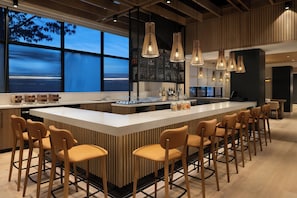 A modern bar area with a central island, bar stools, hanging pendant lights, and a view of the sea through large windows.