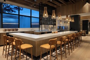 A modern bar area with a central island, bar stools, hanging pendant lights, and a view of the sea through large windows.