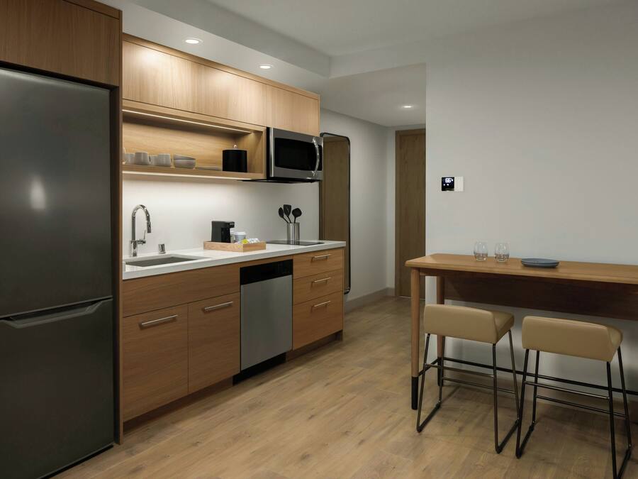 A modern kitchen with wooden cabinets, a stainless steel refrigerator, a microwave, and a sink. There is a wooden dining table with two chairs.