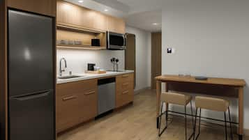 A modern kitchen with wooden cabinets, a stainless steel refrigerator, a microwave, and a sink. There is a wooden dining table with two chairs.
