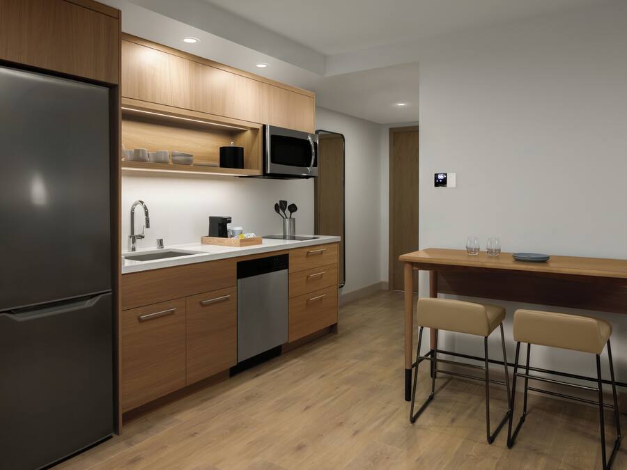 A modern kitchen with wooden cabinets, a stainless steel refrigerator, a microwave, and a sink. There is a wooden dining table with two chairs.