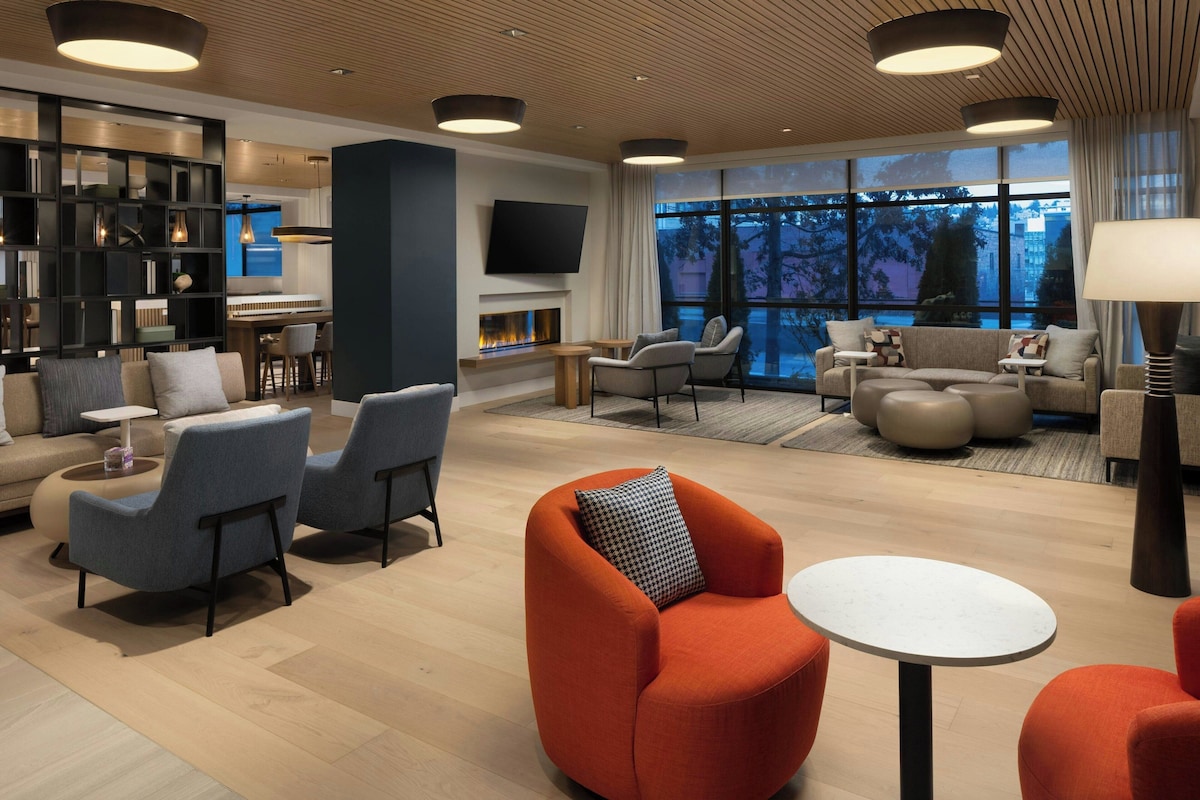A modern hotel lobby with a fireplace, seating areas, and a large window offering a view of the outdoors.