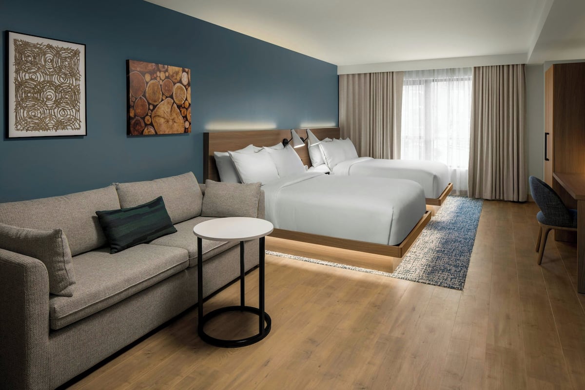 A modern hotel room with a sofa, a round table, a bed with two pillows, and a large window with curtains.