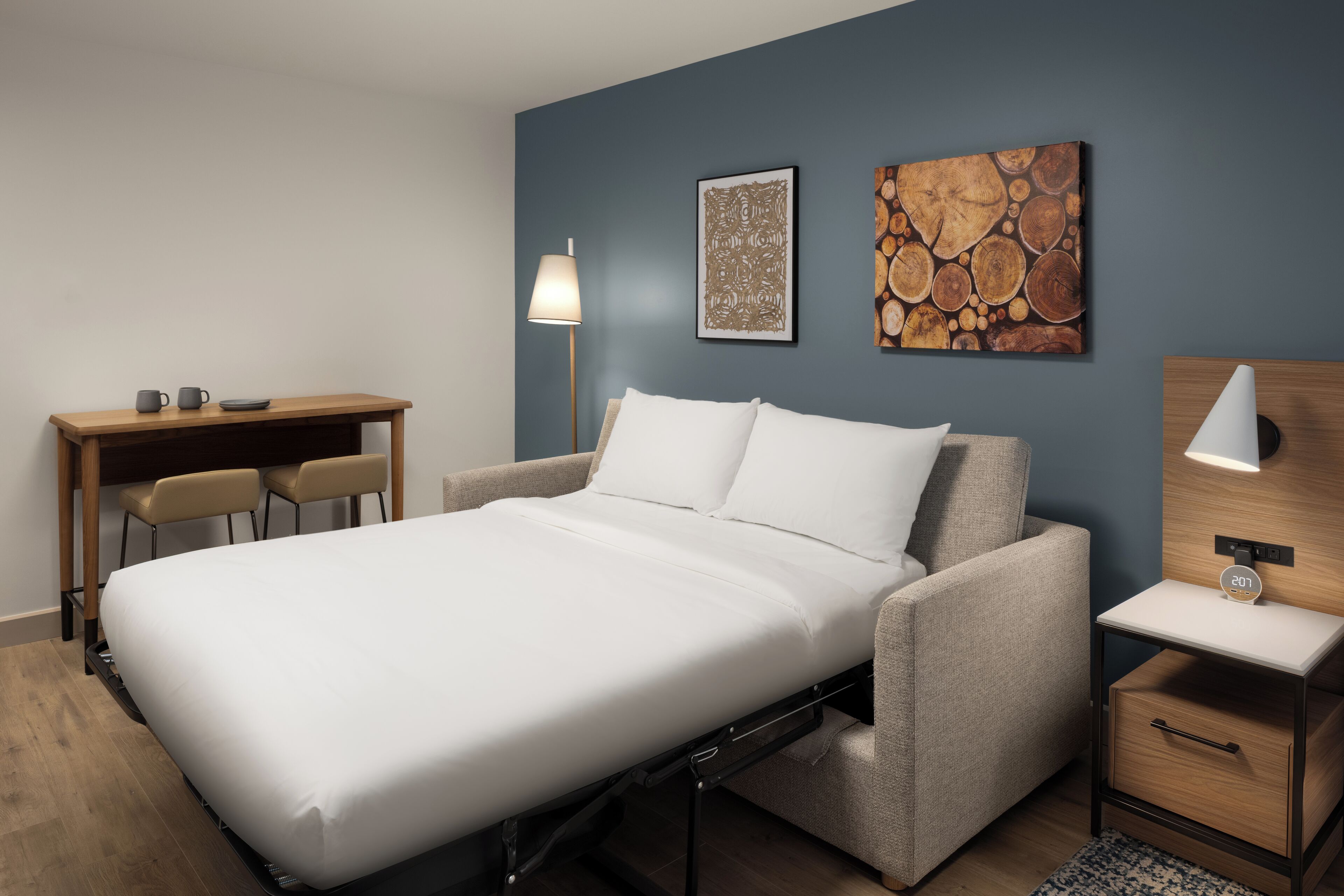 A modern bedroom with a bed, a desk, two chairs, a lamp, and artwork on the wall.