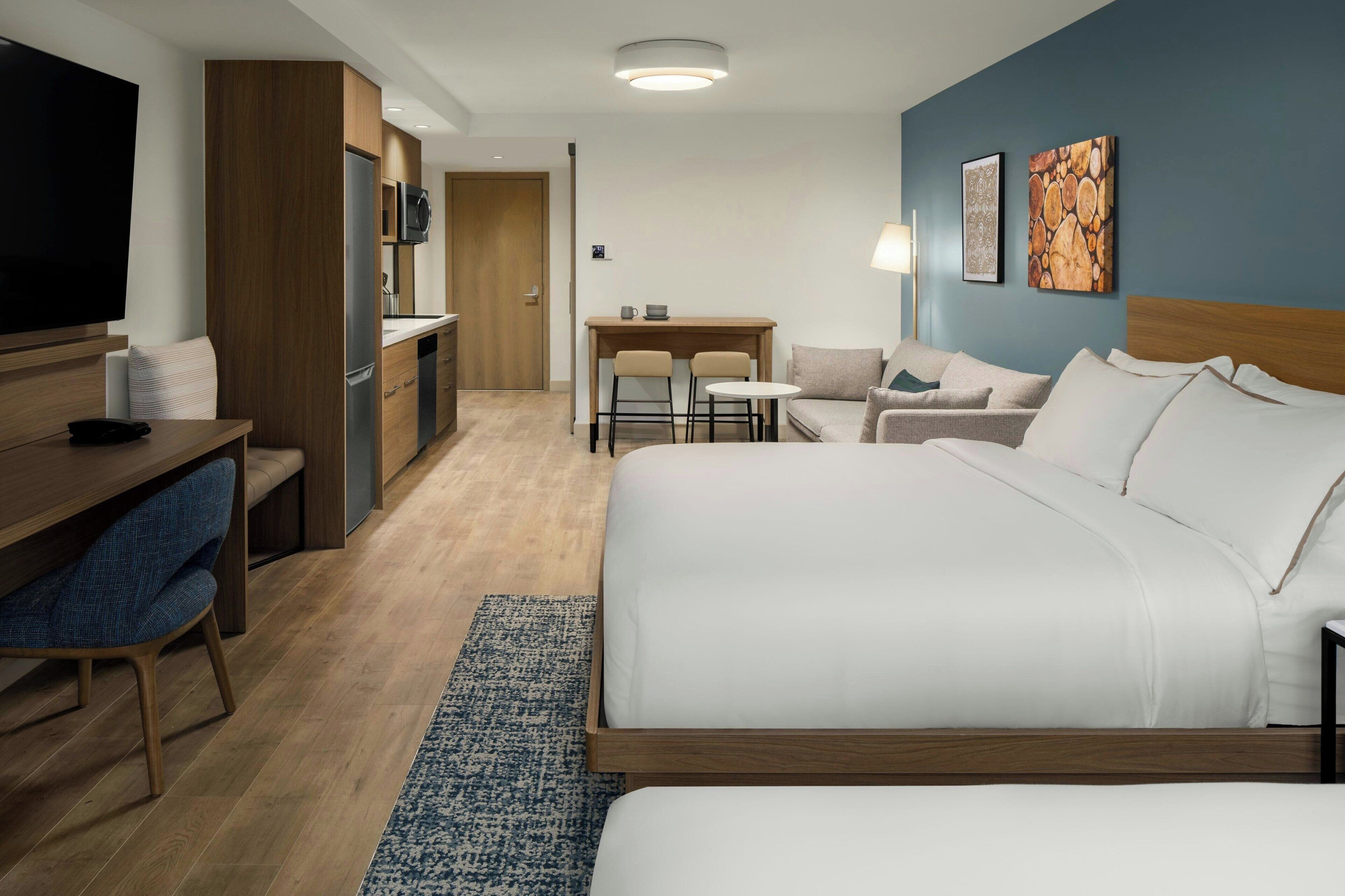 A hotel room with a large bed, a desk with a chair, a small table with two chairs, a TV, and artwork on the wall.