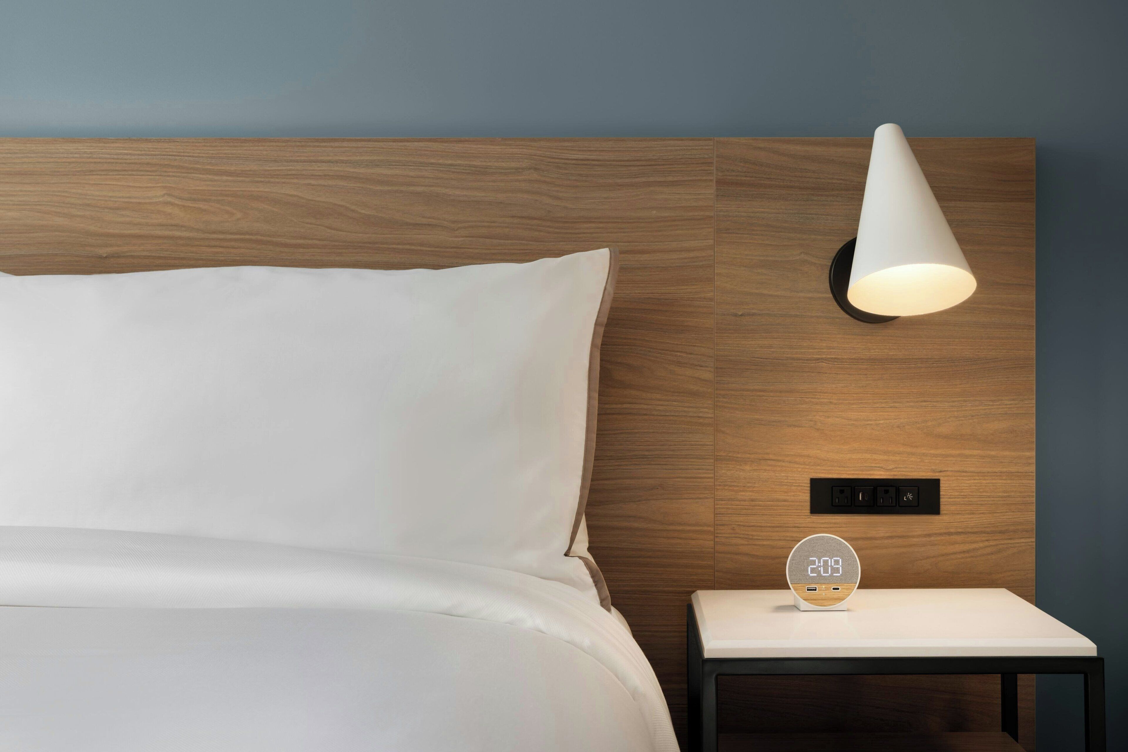 Una habitación de hotel moderna con cabecera de madera, una mesita de noche con un reloj digital y una lámpara de pared.