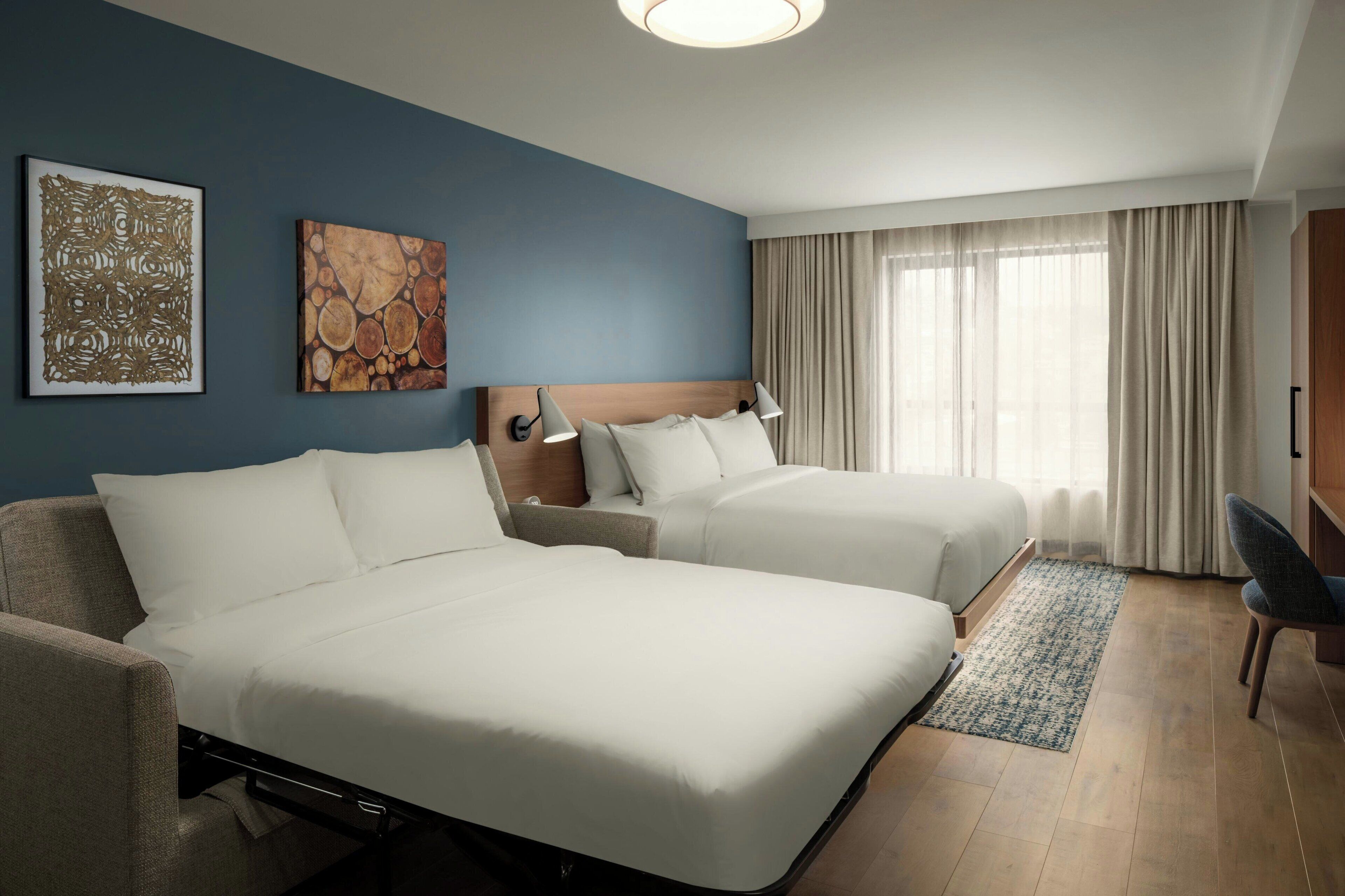 Habitación de hotel con dos camas, un escritorio, una silla, un tapete y obras de arte en las paredes.