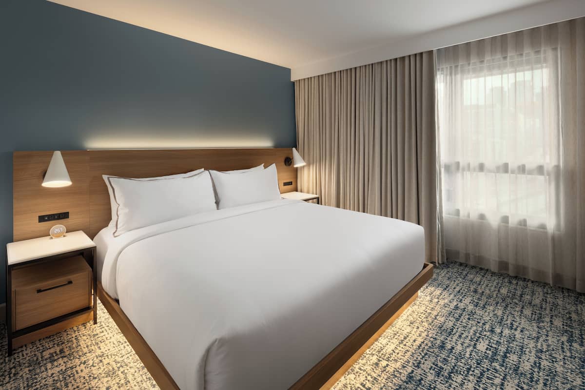 A neatly made bed with a wooden headboard, a bedside table with a lamp, and a large window with sheer curtains.