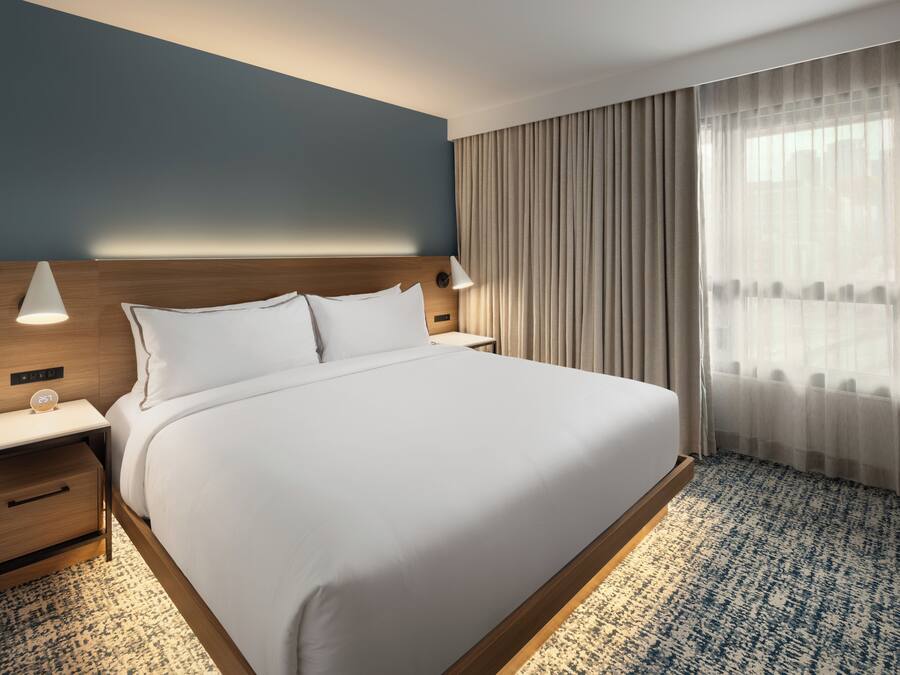 A neatly made bed with a wooden headboard, a bedside table with a lamp, and a large window with sheer curtains.