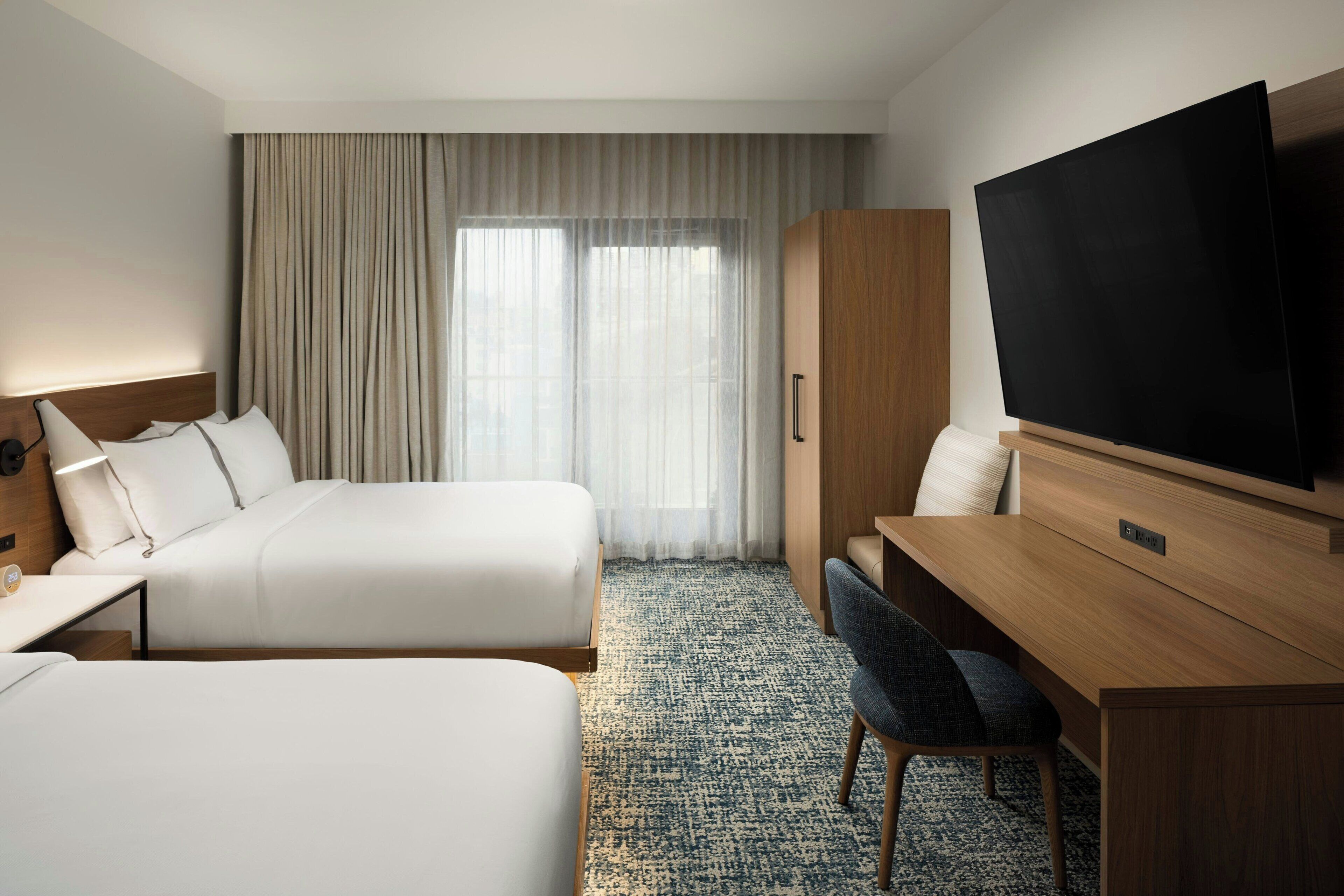 A hotel room with two beds, a desk, a chair, a television, and a large window with curtains.