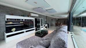 Un salon moderne doté d’un canapé d’angle gris, d’un meuble de rangement intégré pour les appareils multimédias et d’un coin repas avec un mur en verre.