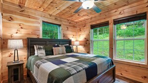 A wooden cabin bedroom with a bed, nightstands, a ceiling fan, and large windows.