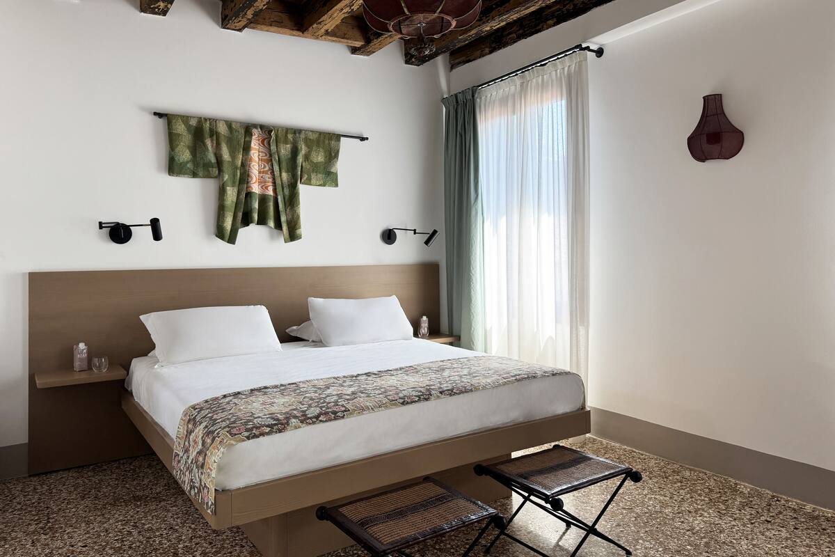 A modern hotel room with a large bed, a small bedside table, a wall-mounted lamp, and a view of the outdoors through sheer curtains.