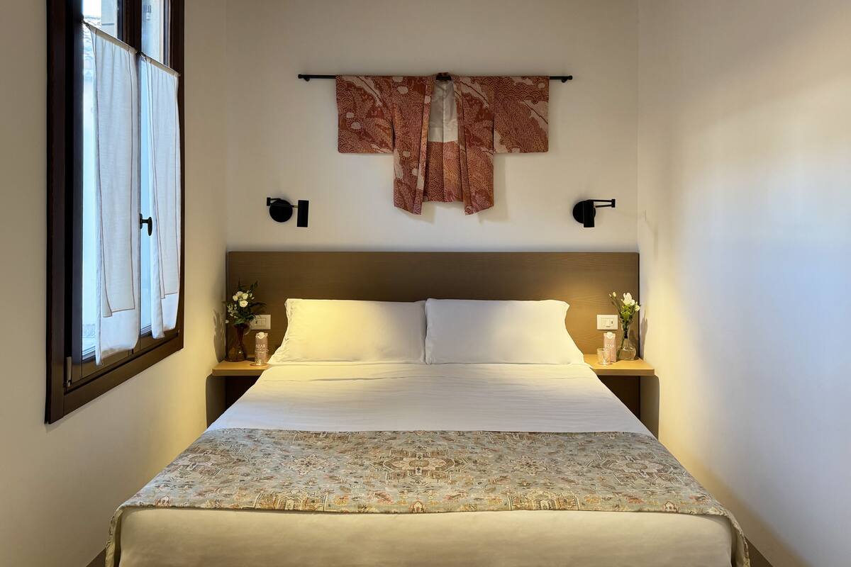 A neatly made bed with a floral bedspread, two bedside tables with lamps, a window with curtains, and a wall-mounted decorative item.