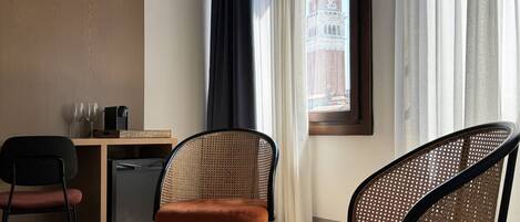 Une chambre d’hôtel avec vue sur une tour, deux fauteuils en osier, une petite table avec un livre et un bureau avec deux verres et une machine à café.