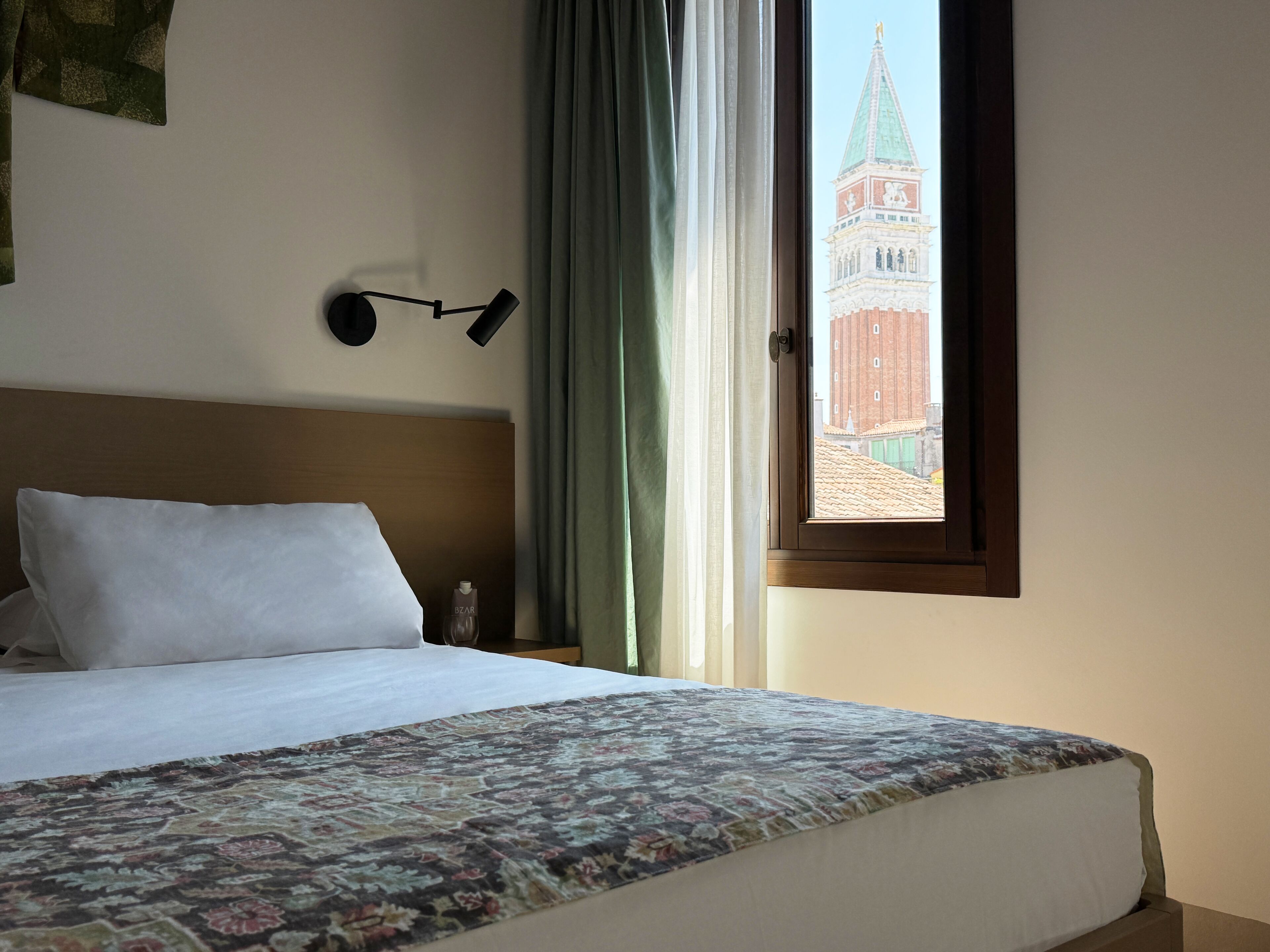 Et hotelværelse med en seng, et natbord, en lampe, et vindue med udsigt til et tårn og et sengetæppe med mønster.