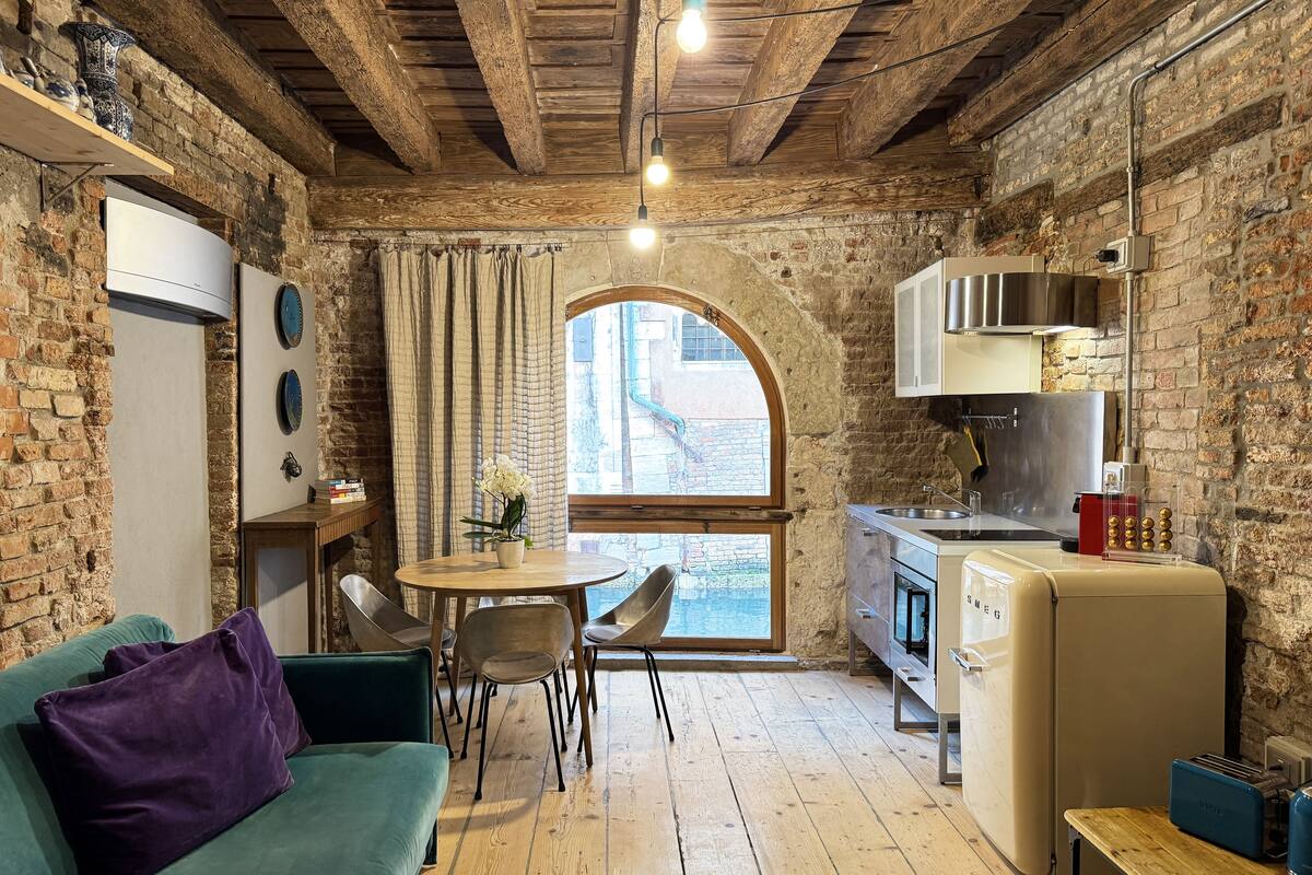 A rustic kitchen with a wooden ceiling, a dining area with a table and chairs, a green sofa, and a window with a view of the outside.