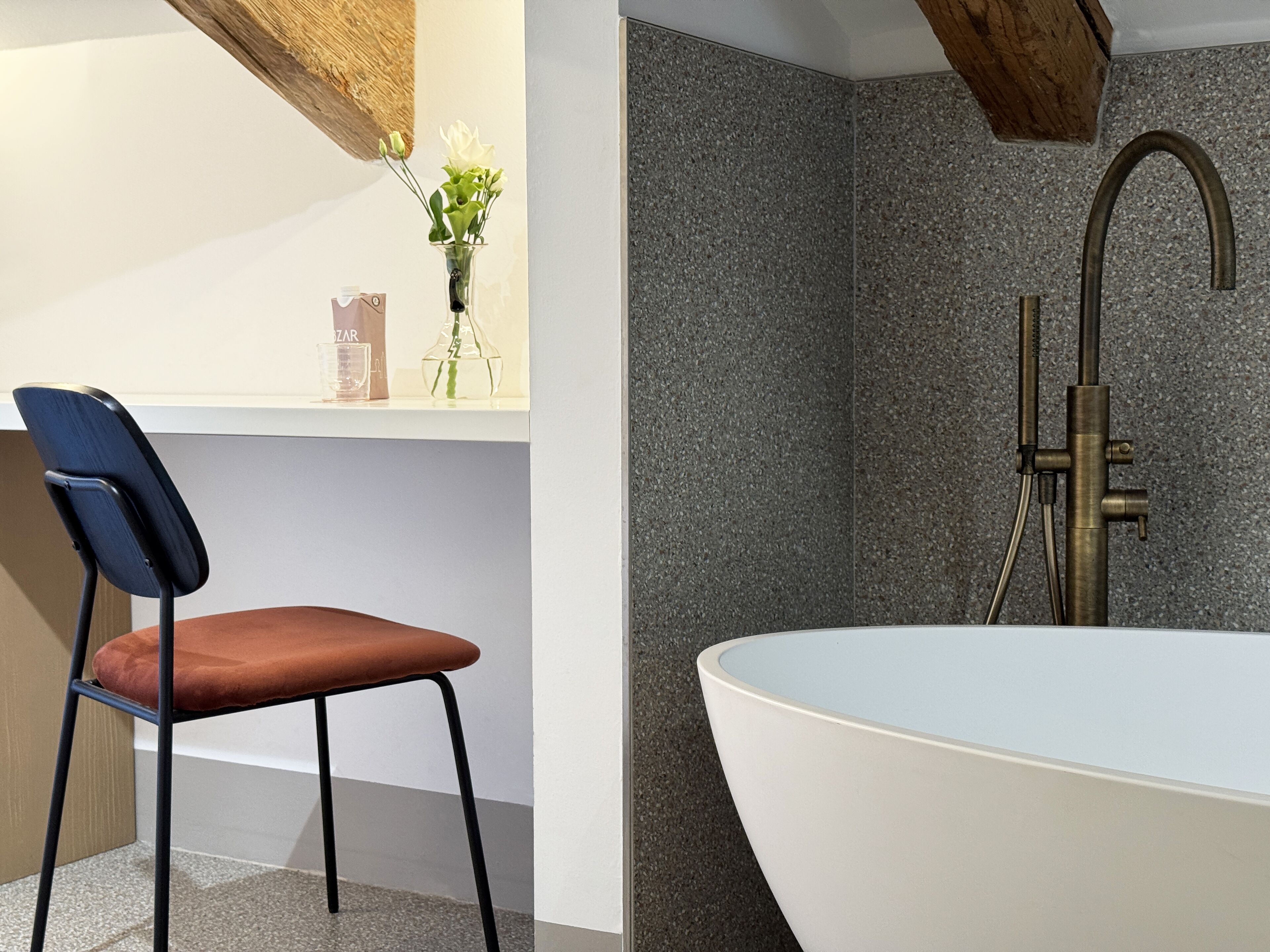 A modern bathroom with a freestanding bathtub, a chair, and a shelf with a vase of flowers.