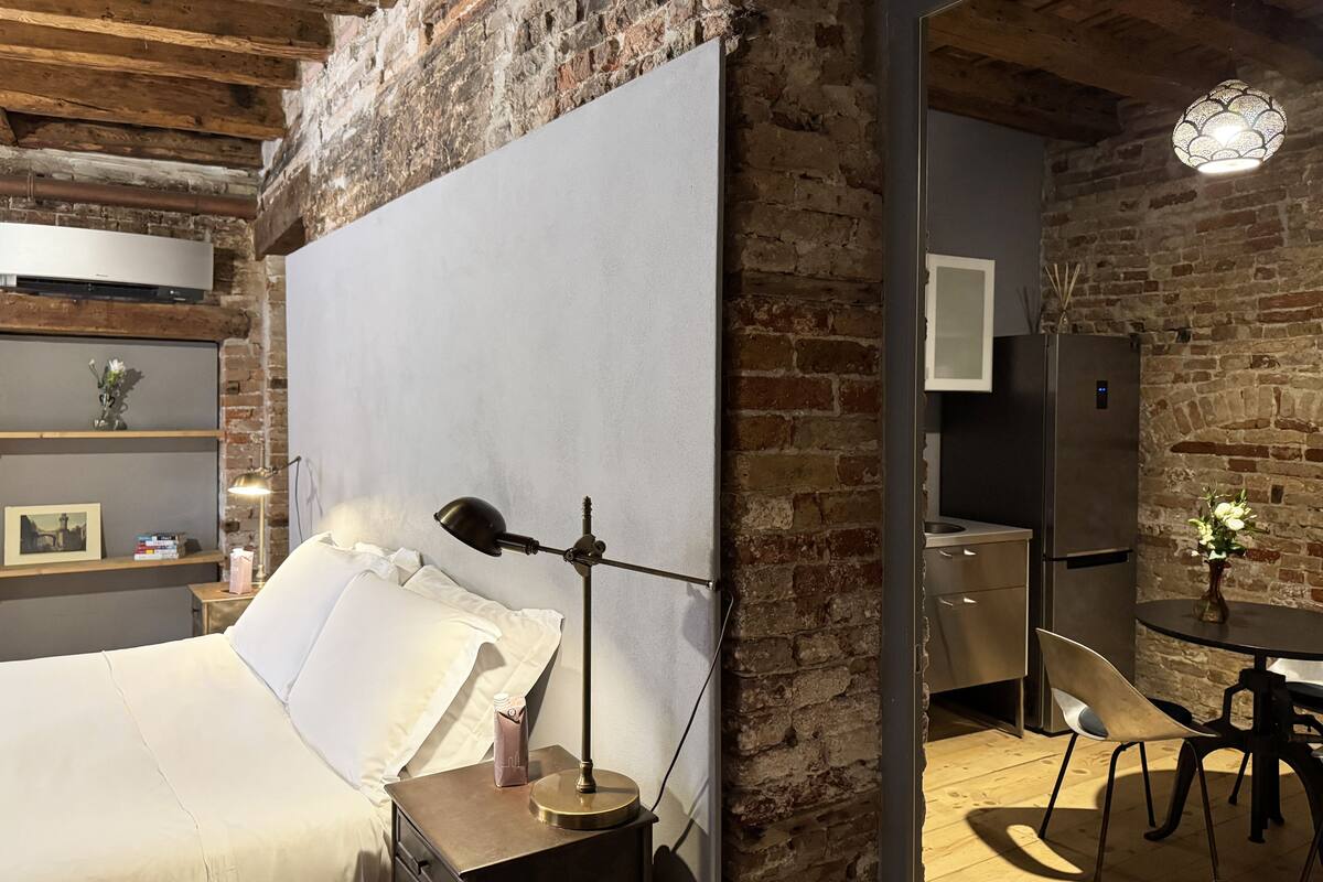 A modern bedroom with a large bed, a bedside table with a lamp, a brick wall, a small kitchen area with a refrigerator, and a dining table with chairs.