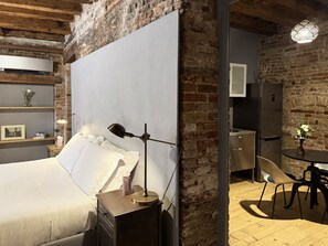 Un dormitorio moderno con una cama grande, una mesita de noche con lámpara, una pared de ladrillo, una pequeña zona de cocina con refrigerador y una mesa de comedor con sillas.